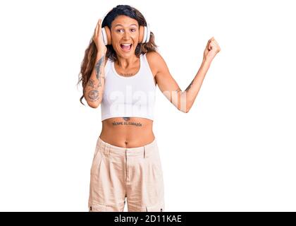 Jeune femme hispanique avec tatouage écoutant de la musique en utilisant des écouteurs criant fier, célébrant la victoire et le succès très excité avec les bras levés Banque D'Images