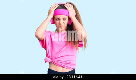 Adorable enfant hispanique fille portant des vêtements de gym et utilisant des écouteurs souffrant de maux de tête désespérés et stressés parce que la douleur et la migraine. Mains sur h Banque D'Images