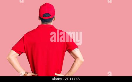 Jeune homme beau avec barbe portant l'uniforme de livraison debout en arrière regarder loin avec les bras sur le corps Banque D'Images