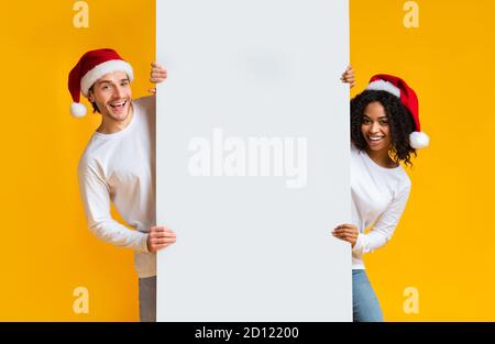 Couple multiracial souriant en chapeaux de père noël tenant un panneau d'affichage blanc pour publicité Banque D'Images