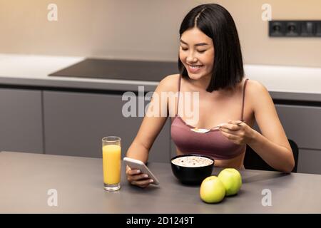 Asian Lady utilisant smartphone Weight-Loss App manger le petit déjeuner dans la cuisine Banque D'Images