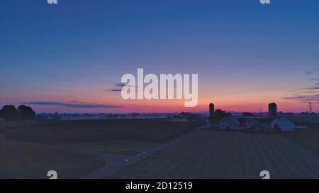 Lever de soleil au-dessus des granges, des silos et des fermes vertes avec des cieux bleus, rouges et orange et quelques nuages avec une brume sur le sol Banque D'Images