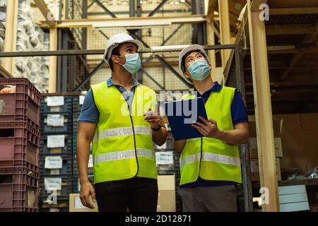 Groupe de jeunes travailleurs d'entrepôt d'usine portant un masque de protection tout en travaillant dans l'industrie logistique intérieure. Des hommes de souche asiatique et indienne vérifient la commande d'articles pendant la pandémie de coronavirus Covid 19 Banque D'Images