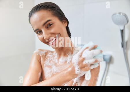 Charmante jeune femme utilisant le loofah exfoliant tout en prenant la douche Banque D'Images