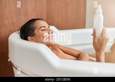 Charmante jeune femme tenant une bouteille de lotion tout en prenant le bain Banque D'Images