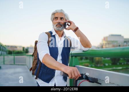 Portrait d'un homme mature avec un scooter électrique en plein air en ville, à l'aide d'un smartphone. Banque D'Images