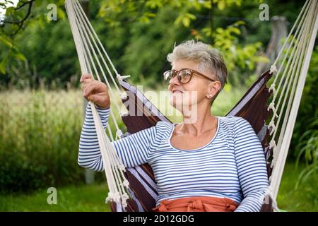 Portrait d'une femme âgée heureuse assise à l'extérieur sur un fauteuil suspendu dans le jardin, reposant. Banque D'Images