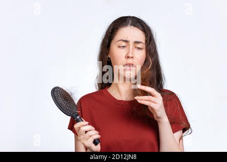 Portrait d'une femme brunette attristée tient un peigne avec les cheveux tombant et regarde les bouts de ses cheveux. Arrière-plan blanc. Concept de perte de cheveux, ba Banque D'Images