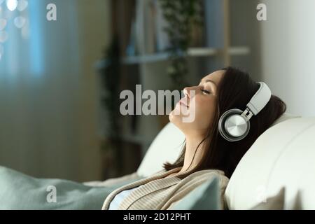 Profil d'une femme détendue écoutant de la musique avec un casque dans la nuit assis sur un canapé dans la vie chambre à la maison Banque D'Images