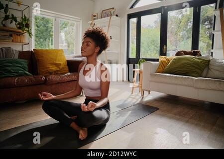 Jeune femme africaine méditant assise en position lotus à la maison sur le tapis de yoga Banque D'Images