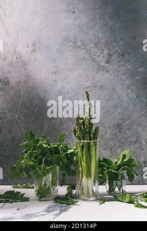 Matières organiques non cuit asperges vertes, botte de coriandre et de menthe fraîche d'herbes dans des bocaux en verre sur la table de cuisine blanche avec texture mur gris comme backgroun Banque D'Images