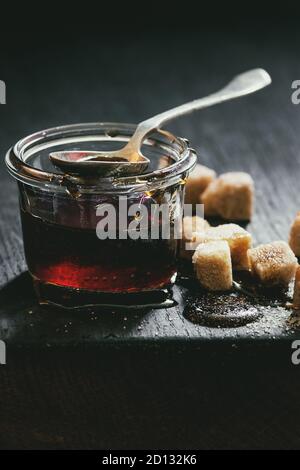 Sucre brun transparent liquide fait maison caramel dans un bocal en verre debout sur planche de bois noir avec cuillère et pouvez cubes de sucre. Close up. La lumière du jour Banque D'Images