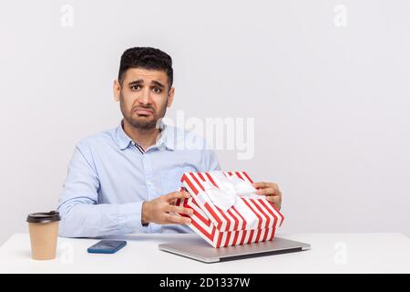 Contrarié homme employé assis dans le bureau lieu de travail, ouvrant la boîte cadeau et regardant la caméra avec l'expression triste déçu, déballer le mauvais présent, celebb Banque D'Images