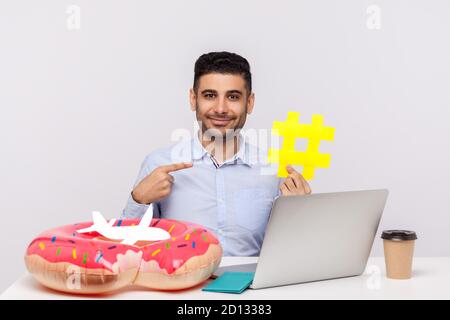 Temps de repos ! Happy man pointant le symbole hashtag, assis sur le lieu de travail du bureau avec un passeport en caoutchouc et un avion en papier sur le bureau. Montrant le signe dièse o Banque D'Images