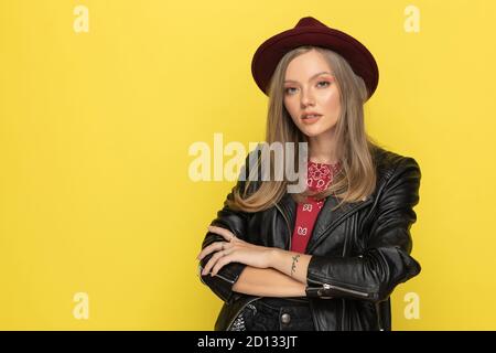 Modèle tendance confiant tenant les mains pliées tout en portant un chapeau et debout sur fond jaune de studio Banque D'Images