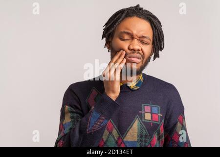 Homme africain barbu frustré avec des dreadlocks touchant la joue et en se réjouissant dans la douleur sensation de terrible douleur dentaire, gingivite, maladie parodontale. Intérieur Banque D'Images