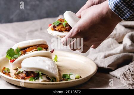 Tenir les mains de l'homme sandwich asiatique brioches bao gua à la vapeur avec du porc, du ventre et des légumes verts servi dans une plaque en céramique sur table avec nappe en lin. Comme Banque D'Images