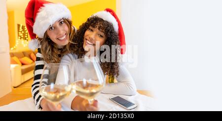 Deux jeunes femmes fines de race blanche et hispanique mixtes à maison regardant l'appareil photo pour griller avec un verre De vin en appel vidéo - Nouveau Holi normal Banque D'Images