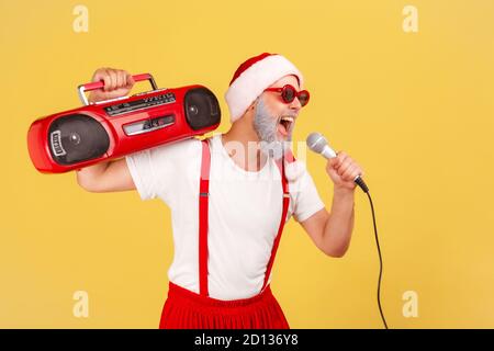 Homme âgé positif et actif dans le chapeau du père noël et lunettes de soleil tenant un magnétophone sur l'épaule chantant des chansons au microphone, karaoké de vacances. Indo Banque D'Images