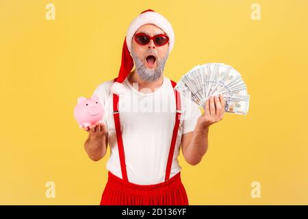 Homme adulte extrêmement choqué dans des lunettes de soleil élégantes et chapeau de père noël tenant la banque de porc et des billets de dollars regardant l'appareil photo, dépôt, cashback. Po Banque D'Images