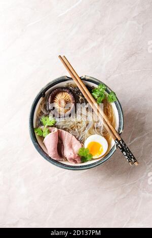Soupe japonaise traditionnelle de nouilles aux champignons shiitake, œufs, tranches de bœuf et légumes verts, servie dans un bol en céramique avec baguettes en bois sur une bande de marbre rose Banque D'Images