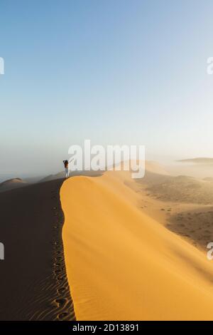Femme marchant au sommet d'une immense dune de sable dans le désert du Sahara marocain. Belle lumière chaude du soleil et brume le matin. Vue de derrière. Concept de liberté. Banque D'Images