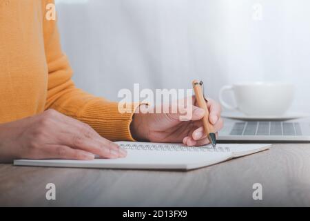 Image rognée d'une femme d'affaires assise à une table et prenant des notes dans un carnet, sur une table est un ordinateur portable et une tasse de café. Banque D'Images