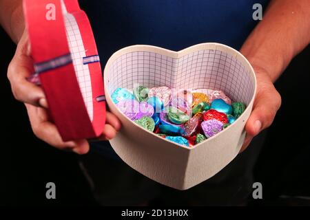 gros plan de la boîte cadeau en forme de coeur de la main de l'homme avec des bonbons isolés sur le noir Banque D'Images