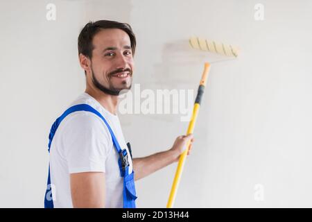 Un repeint barbu en vêtements de travail peint le mur avec un rouleau. Il sourit amical et pose pour la caméra Banque D'Images