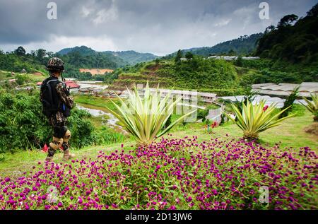 Vue arrière d'un soldat des forces spéciales asiatiques en uniforme de camouflage, casque protégé avec arme à feu dans les mains lors d'une patrouille de sécurité au-delà du jardin fleuri. Banque D'Images