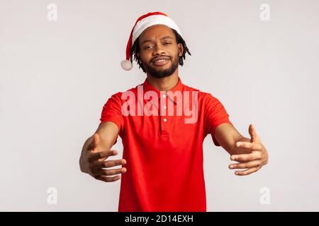 Sympathique homme barbu afro-américain avec des dreadlocks dans le chapeau du père noël tenant les mains prêtes à embrasser, câlins libres le soir de noël. Prise de vue en studio Banque D'Images