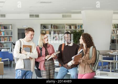 Portrait d'un groupe multiethnique d'étudiants se tenant dans la bibliothèque de l'université et discutant tout en tenant des livres et des sacs à dos, espace de copie ci-dessus Banque D'Images