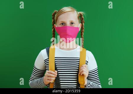 La vie durant la pandémie de Covid-19. Portrait d'une jeune fille d'école moderne en blouse blanche avec sac à dos jaune et masque rose isolé sur tableau noir vert backgrou Banque D'Images