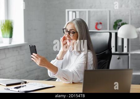 Femme sénior aux cheveux gris qui utilise un smartphone et qui sourit. Bonne femme d'affaires utilisant des applications de téléphone mobile, des messages texte, la navigation sur Internet Banque D'Images