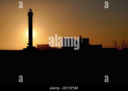 Alors que le soleil se dépare derrière le phare de 43 m (141 pi) qui a alerté la navigation de la Manche en naviguant dans le détroit de Douvres au risque de la pointe de bardeaux Dungeness qui jouvrait dans la mer depuis son ouverture en 1961, il semble éclipser un phare édouardien antérieur, ouvert en 1904, qui utilisait une lampe à paraffine. Les réacteurs et la salle à turbine de Dungeness A, une centrale nucléaire de Magnox qui a produit de l'électricité de 1965 à 2006, ont également été mis hors service en toute sécurité. Banque D'Images