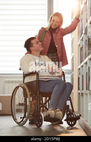 Portrait vertical complet du jeune homme en fauteuil roulant vers l'intérieur école avec une amie l'aidant dans la bibliothèque éclairée par lumière du soleil Banque D'Images