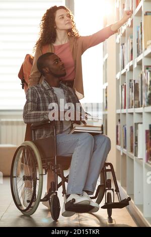 Portrait vertical complet du jeune homme afro-américain en fauteuil roulant à l'école avec une amie qui l'aide dans la bibliothèque éclairée par la lumière du soleil Banque D'Images