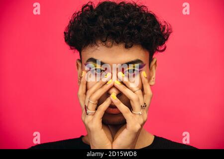 Homme liquide de sexe portant l'ombre des yeux de couleur arc-en-ciel et le visage smiley sur l'ongle. Mâle transgenre avec maquillage funky sur fond rouge. Banque D'Images