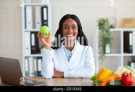 Diététicienne noire positive tenant des fruits de pomme et regardant caméra en clinique Banque D'Images