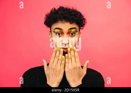 Homme gay portant l'ombre des yeux de couleur arc-en-ciel et le visage souriant sur l'ongle sur fond rouge. Homme transgenre avec maquillage funky. Banque D'Images