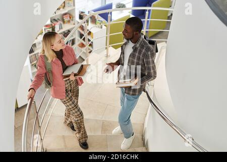 Portrait à grand angle de deux étudiants discutant tout en se tenant sur l'escalier en spirale dans l'université et tenant des livres, l'espace de copie Banque D'Images