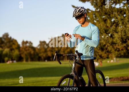 Vérification des résultats. Jeune homme sportif, cycliste professionnel en vêtements de sport et casque de protection debout avec son vélo dans le parc et en utilisant son smartphone. Style de vie actif et sport Banque D'Images