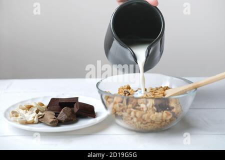 granola maison au chocolat et aux noix. granola dans un bol transparent avec versement de lait et de chocolat sur une assiette. noix de cajou et morceaux de cacao crus. concept petit déjeuner biologique. Banque D'Images