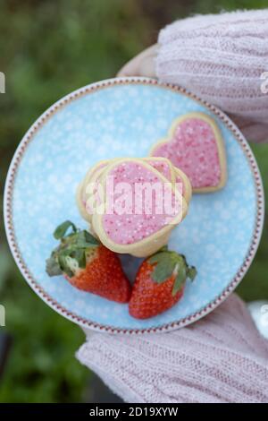 Fille tenant une assiette avec des biscuits en forme de coeur rose accompagnés par les fraises Banque D'Images