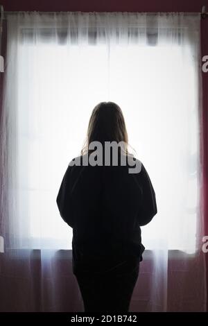 Silhouette d'une jeune femme qui donne sur une fenêtre avec des rideaux blancs transparents. Banque D'Images