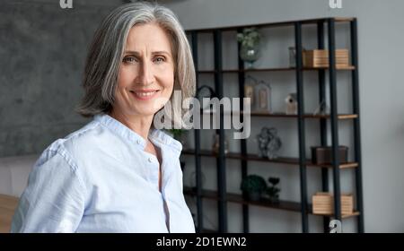 Souriante confiante femme d'âge moyen aux cheveux gris debout au bureau, portrait. Banque D'Images