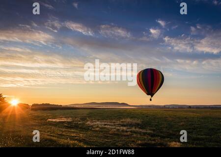 Le ballon d'air chaud dans des bandes arc-en-ciel colorées commence l'ascension champ de ferme au soleil se lève ciel bleu ciel nuageux Banque D'Images