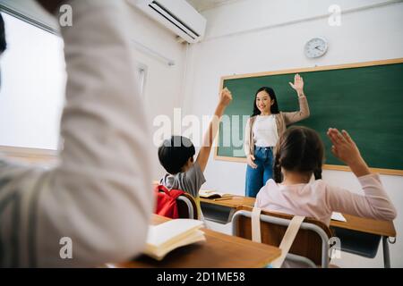 Professeur d'école asiatique avec des élèves qui élèvent la main. Jeune femme travaillant à l'école avec le bras levé, les enfants de l'école mettant leurs mains vers le haut pour répondre à la question, enthousiasme, désireux, plaisir. Banque D'Images