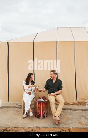 Jeune couple heureux appréciant le thé à la menthe marocain traditionnel dans le camp de glamping dans le désert du Sahara, au Maroc. Banque D'Images