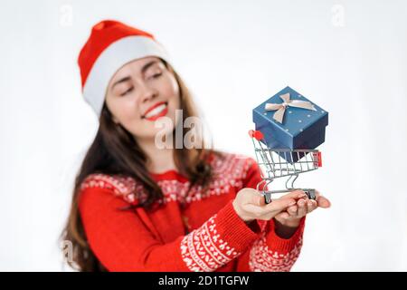 Portrait d'une jeune femme souriante dans un chandail du nouvel an et chapeau de père Noël, tenant une mini-voiturette avec un cadeau. Arrière-plan blanc. Le concept de la Christm Banque D'Images